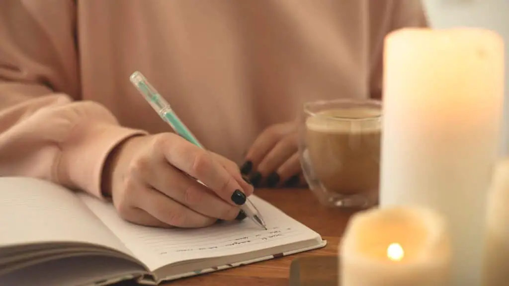 woman writing a prayer list at a table with coffee and a candle