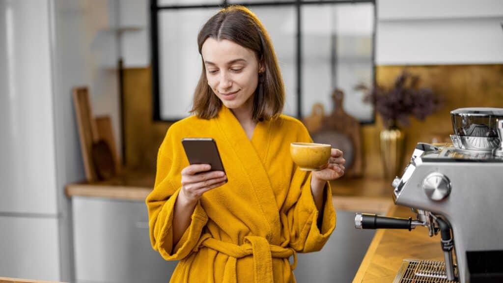 woman wearing a yellow robe and drinking coffee while checking her phone