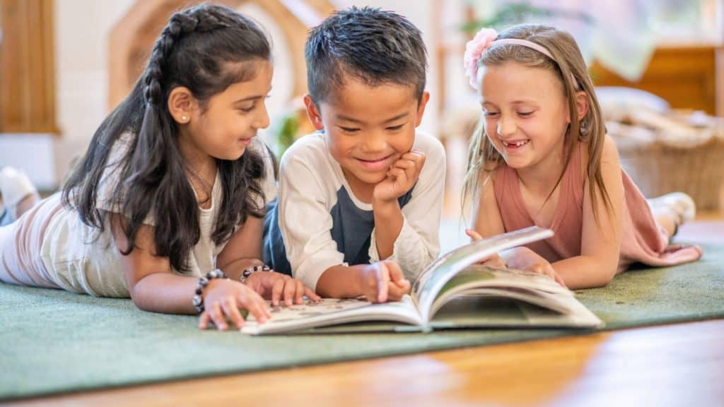 three children reading a book together while lying on the floor