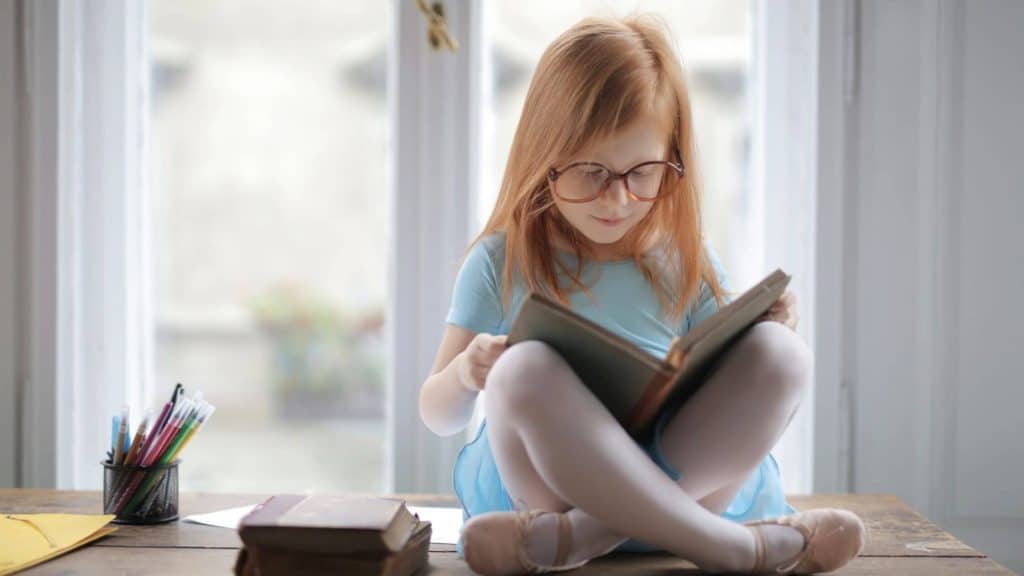 little girl with big glasses reading in front of a window