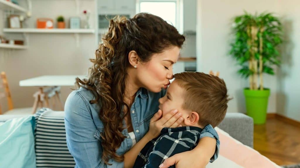 woman with curly hair holding her small son and kissing his forehead 