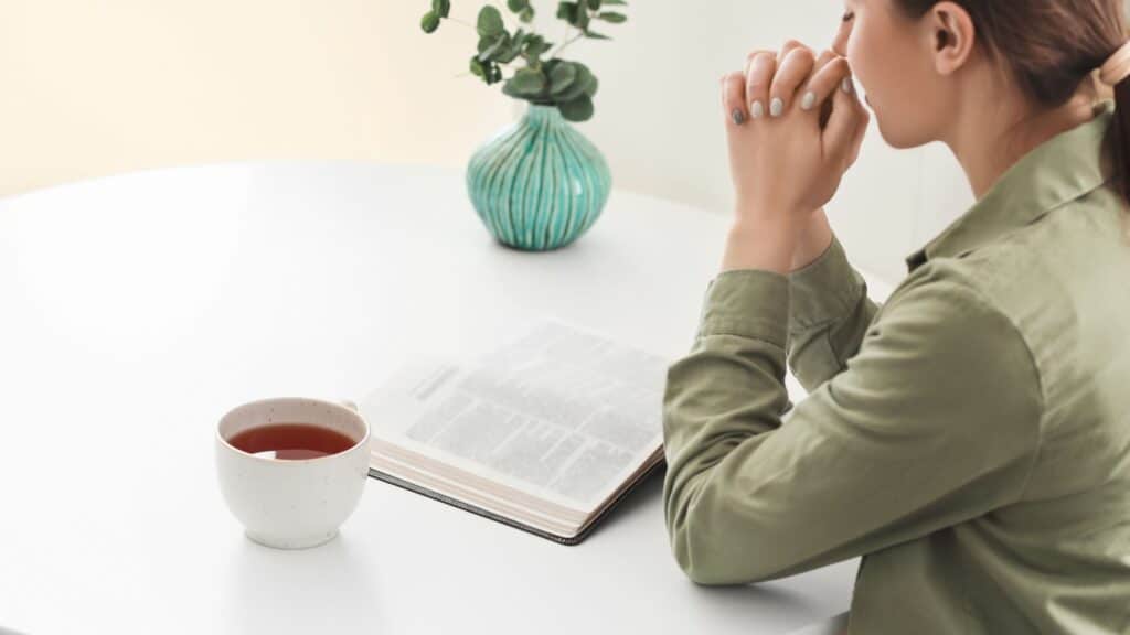woman with hands clasped in prayer over her Bible on a table with a plant and coffee