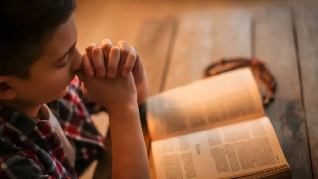 young boy holding a Bible on his lap with hands clasped in prayer