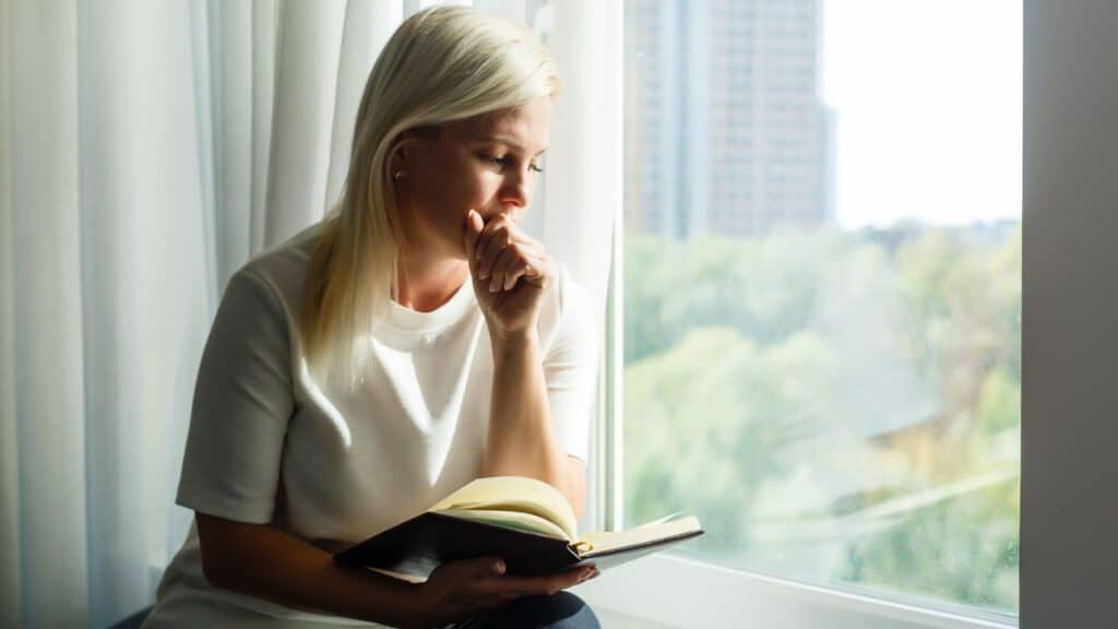 woman praying in front of a window while holding her Bible