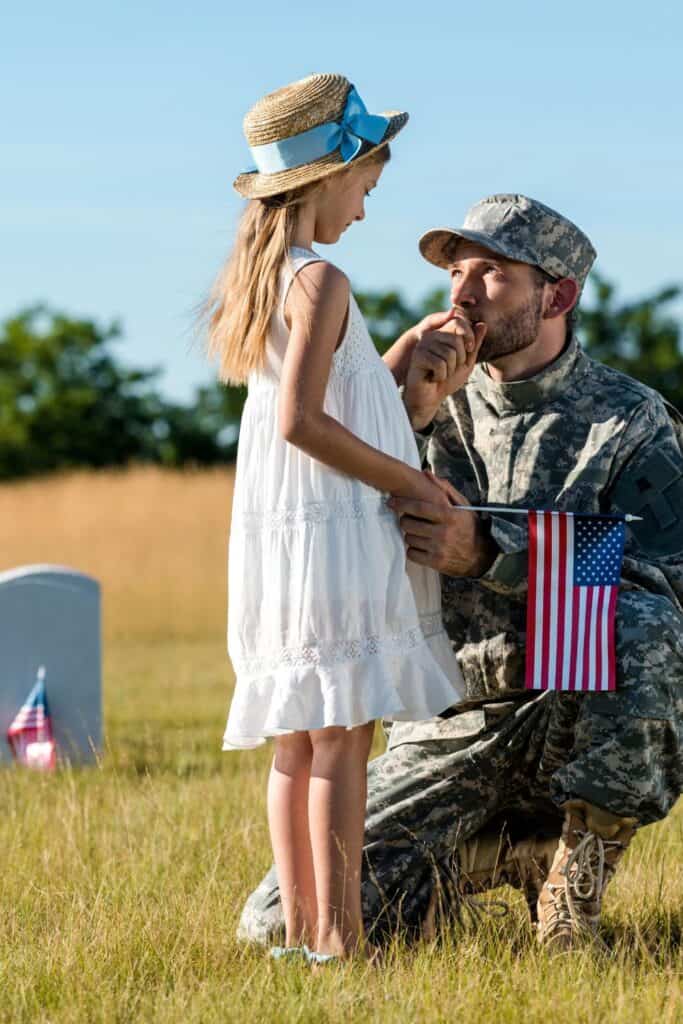 man in military uniform kneeling down to kiss the hand of a small girl wearing a sundress and hat with a decorated military grave in the background