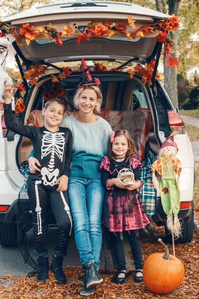 mother leaning against a car decorated with fall foliage with two children dressed in costumes