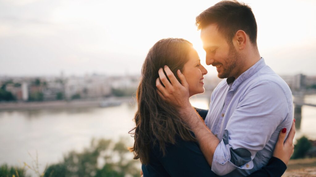 smiling husband and wife standing beside a lake at sunset