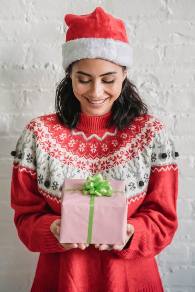 woman wearing a Christmas sweater and a Santa hat and holding a gift